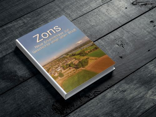 Ein geschlossenes Buch auf einem Tisch, mit einem einfachen Cover-Design. Der Titel "Zons. Neue Erkenntnisse über die Geschichte einer alten Stadt" ist deutlich sichtbar und das Buch scheint in gutem Zustand zu sein, bereit zum Lesen oder zur Aufbewahrung in einer Bibliothek.